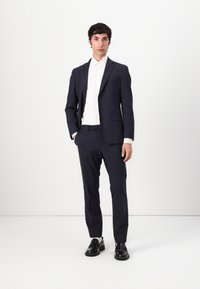 Traje azul marino con chaqueta a medida y pantalones de frente plano, combinado con una camisa blanca. Cuenta con un corte ajustado y una textura suave. Zapatos negros completan el look.