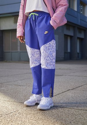 Osoba ubrana w niebiesko-białe spodnie Nike w wzory, białe sneakersy, biały krótki top i różową puchową kurtkę stojąca na zewnątrz.