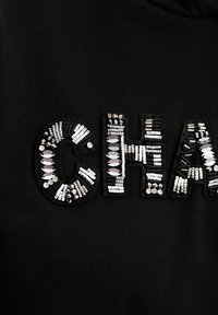 Czarna bluza z dużymi literami "CH" ozdobionymi różnorodnymi koralikami i cekinami w odcieniach srebra, bieli i czerni.