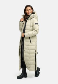 Lang lys beige dunjakke med hætte, featuring zip lukning, quiltet tekstur og sorte detaljer. Båret over mørkt tøj og støvler.