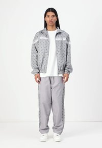 Grijze geprinte jas met witte accenten en bijpassende grijze broek; beide hebben een ruitpatroon en een losse pasvorm, gecombineerd met witte sneakers.