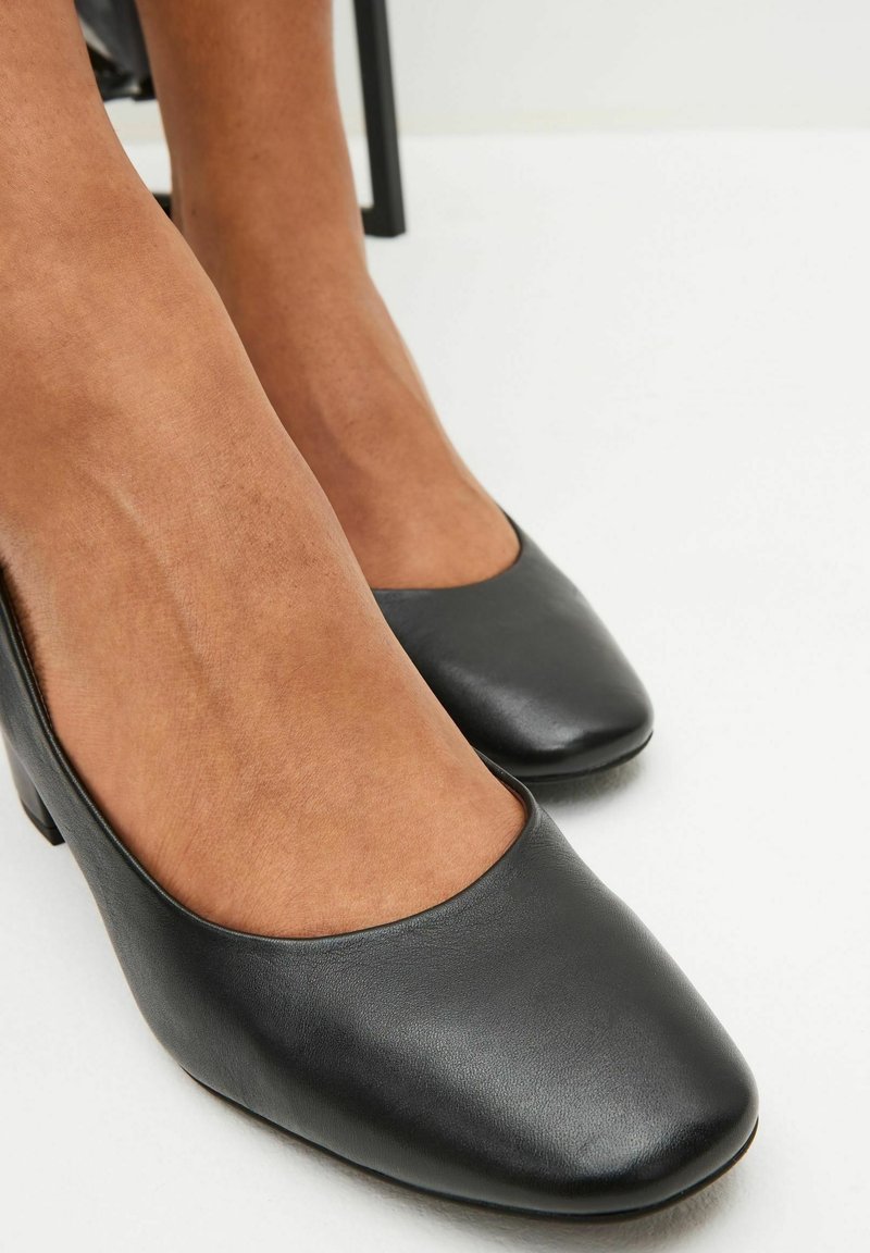 Pieds portant des chaussures en cuir noires à talons bas avec des bouts carrés arrondis sur un sol blanc, montrant une vue de face et de profil.