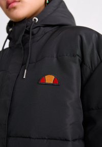 Padded sort jakke med hætte, der har et farverigt broderet logo i rød, orange og gul på brystet. Glat tekstur, holdbart materiale.