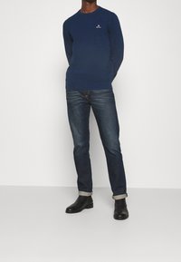 Marineblauer Langarmpullover mit Rundhals, versehen mit einem kleinen weißen Logo. Kombiniert mit dunklen Jeans und schwarzen Stiefeln, die Ärmel hochgekrempelt.