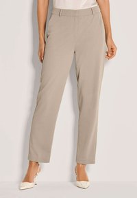 Vrouw in beige straight-leg broek en witte slingback hakken met spitse neus, staand tegen een eenvoudige witte achtergrond.