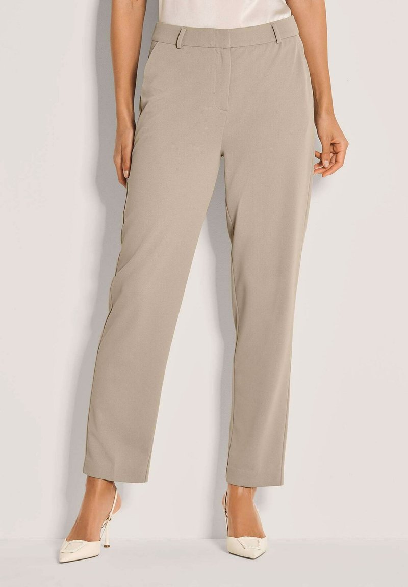 Vrouw in beige straight-leg broek en witte slingback hakken met spitse neus, staand tegen een eenvoudige witte achtergrond.