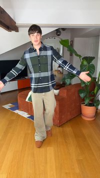 Junger Mann mit kariertem Hemd und beigen Hosen steht mit offenen Armen in einem Wohnzimmer mit Holzboden, orangefarbenem Sofa, Pflanzen und Fernseher.