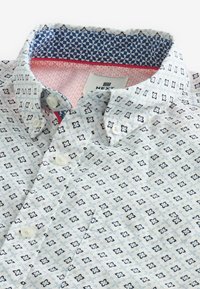 Camisa blanca abotonada con pequeños estampados florales geométricos en azul marino y tela con un patrón contrastante en azul y rosa en el interior del cuello.