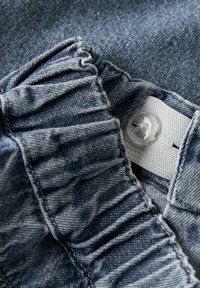 Džinsa audums ar savākto tekstūru, attēlo pogas aizdari un balto birku. Krāsa ir vidēji zila ar smalku izbalējušu rakstu.