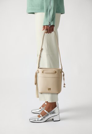 Sac seau en cuir beige avec des ferrures en or, doté d'un dessus à cordon coulissant, d'une texture lisse et d'une bandoulière amovible. Chaussures à talons bloc argentés.