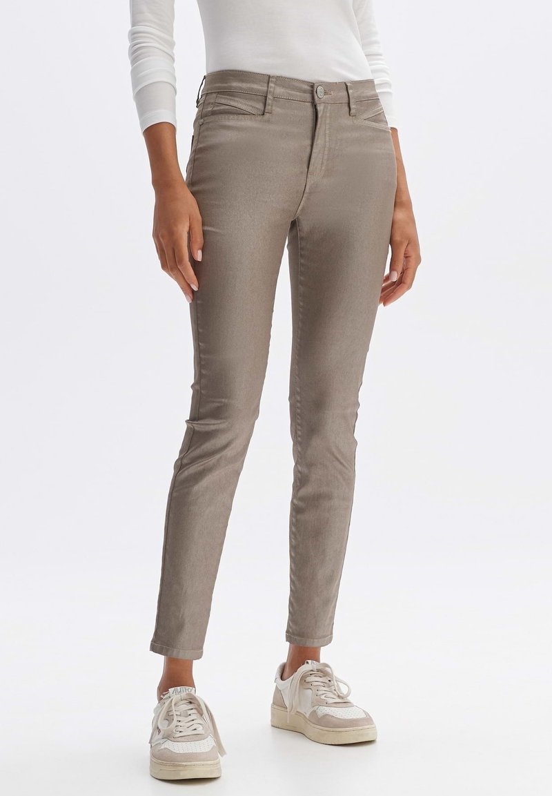 Pantalon slim taupe porté avec un haut à manches longues blanc et des baskets écrues, montré de la taille aux pieds sur un fond uni.
