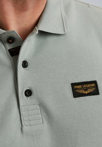 Světle zelená polo tričko s texturovanou tkaninou, černými knoflíky a nášivkou s nápisem "PME LEGEND" s křídly.