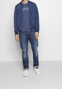 Giacca bomber navy con polsini a costine, indossata sopra una maglietta grigia con logo rosso e bianco, abbinata a jeans blu e sneakers bianche.
