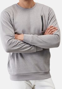 Uomo che indossa un felpa grigia chiara a maniche lunghe con tasca con zip sul petto, braccia incrociate, abbinata a pantaloni bianchi.