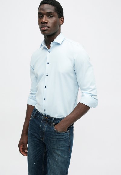 Ανοιχτό μπλε πουκάμισο με μακριά μανίκια και κανονικό γιακά, με ναυτικές κουμπιά, σε συνδυασμό με σκούρα μπλε τζιν με υφή τζιν και διακριτικές λεπτομέρειες.