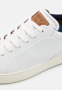 Vita lädersneakers med texturerad sidopanel, brun konstläderetikett och vita snören. Gummisula med sömdetalj runt kanten.