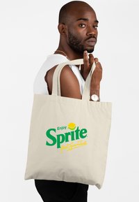 Beżowa torba na zakupy z bawełny z zielonym logo "Sprite" i �żółtymi akcentami, trzymana przez osobę z krótkimi włosami, ubraną w bezrękawnik biały.