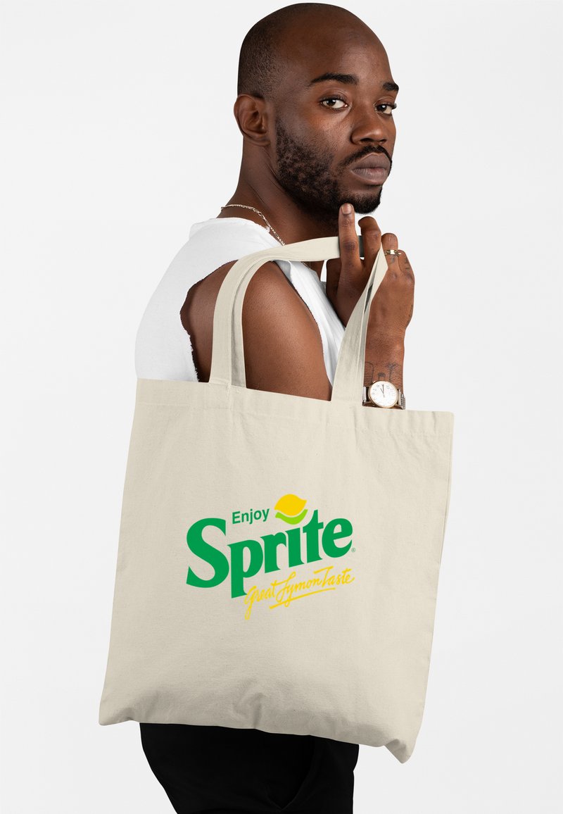 Beżowa torba na zakupy z bawełny z zielonym logo "Sprite" i żółtymi akcentami, trzymana przez osobę z krótkimi włosami, ubraną w bezrękawnik biały.