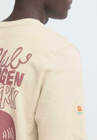 Beige katoenen t-shirt met bordeauxrode bedrukt tekst en een logolabel op de mouw. Korte mouwen en een ronde hals.