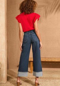 Punainen lyhythihainen paita, joka on yhdistetty korkeavyötäröisiin, leveälahkeisiin sinisiin farkkuihin, joissa on käänteet ja kaksi takataskua. Neutraali tausta.