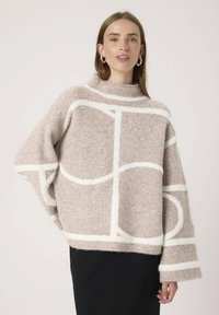 Beige uldtrøje med høj hals, oversized pasform og hvide geometriske mønstre. Blødt, struktureret stof og brede ærmer.