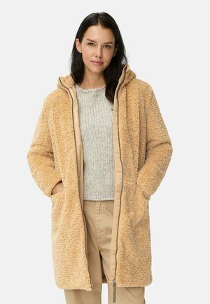Tan nepbontjas met capuchon, voorzien van zijzakken, een zachte textuur en contrasterende beige voering; gedragen over een lichtgrijze trui.