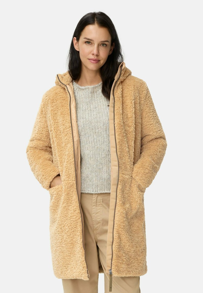 Tan nepbontjas met capuchon, voorzien van zijzakken, een zachte textuur en contrasterende beige voering; gedragen over een lichtgrijze trui.
