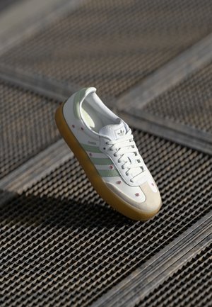 Vit texturerad sneaker med gröna ränder, krämiga accenter och rosa blommig broderi. Har en gummisula och rundad tådesign.