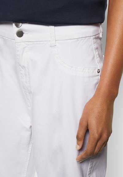 Calças de ganga brancas com dois botões prateados na cintura, bolsos laterais e uma textura suave. Uma mão descansa na lateral da perna da calça.