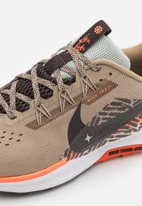 Nike trail løbesko i beige og brun, med mesh-overdel, strukturerede overlays, orange accenter og et sort Swoosh-logo.