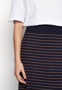 Personne portant une chemise blanche à manches courtes et une jupe bleu marine avec de fines rayures horizontales beige et une ceinture côtelée.