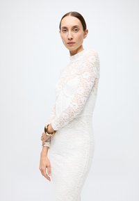 White lace dress with long sleeves, high neck, fitted silhouette. Features floral patterns and a smooth texture, paired with a gold bangle.
