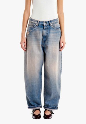 Donna che indossa jeans blu slavati e larghi con orli arrotolati, una canottiera bianca senza maniche e ballerine scure con cinturini alla caviglia.