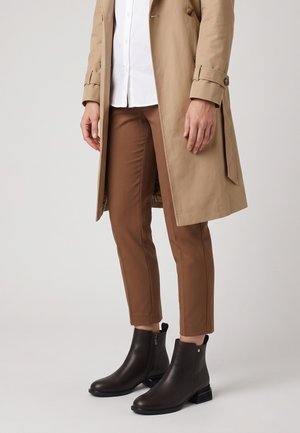 Person trägt braune Stiefeletten, braune enganliegende Hose, weißes Hemd und einen beigen Trenchcoat und steht vor einem schlichten Hintergrund.