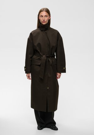 Femme aux cheveux raides portant un long manteau sombre avec un col montant, une ceinture, de grandes poches et des manches larges, debout sur un fond uni.
