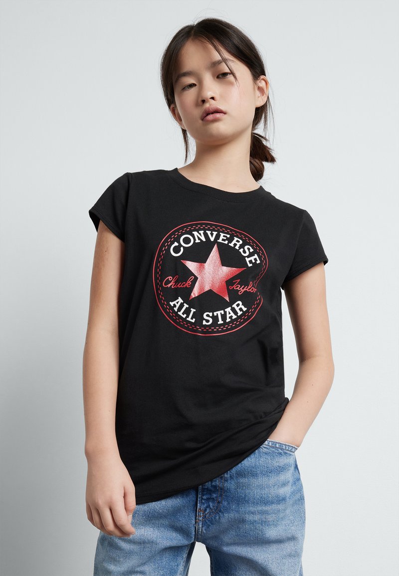 Melna kokvilnas T-krekls ar sarkanu un baltu apļveida grafiku, kurā ir zvaigzne un teksts "CONVERSE ALL STAR", apvienots ar gaiši ziliem džinsiem.