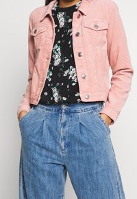 Lyserød corduroy jakke med sølvknapper over en sort blomsterbluse, parret med højtaljede blå denimbukser med folder.