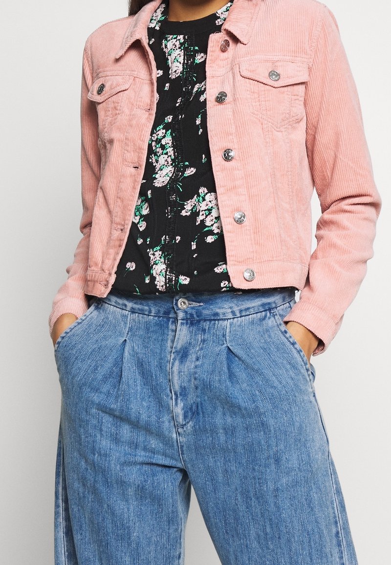 Lyserød corduroy jakke med sølvknapper over en sort blomsterbluse, parret med højtaljede blå denimbukser med folder.