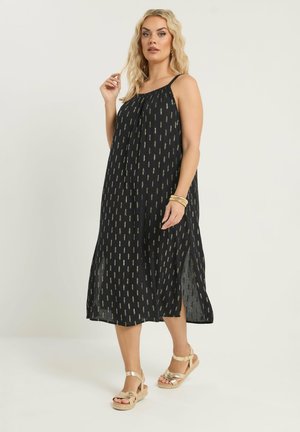 Blondynka w czarnej, bezrękawnikowej sukience midi z złotym wzorem i złotych sandałach, pozująca z dłonią przy włosach oraz bransoletkami na nadgarstku.