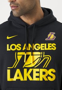 Hoodie preto com texto e logotipo "Los Angeles Lakers" em amarelo. Apresenta o símbolo da Nike em amarelo. Cordões com pontas amarelas, textura suave.