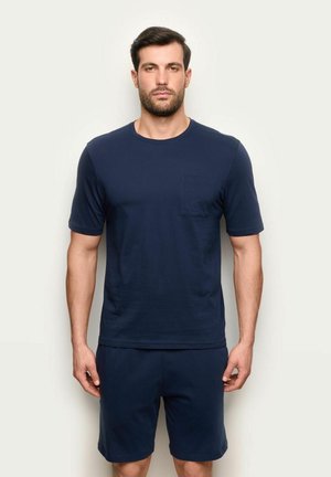 Mann steht vor schlichter Hintergrund, trägt ein dunkelblaues kurzärmeliges Hemd mit Brusttasche und passende Shorts, schaut nach vorne mit neutralem Gesichtsausdruck.