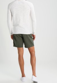 Maglietta grigia chiara a maniche lunghe con tessuto testurizzato, abbinata a pantaloni corti verdi scuri e sneaker bianche, vista da dietro.
