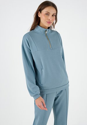 Felpa leggera azzurro con zip a un quarto e colletto alto, tasca frontale e tessuto testurizzato. Abbinata a pantaloni leggeri azzurri.