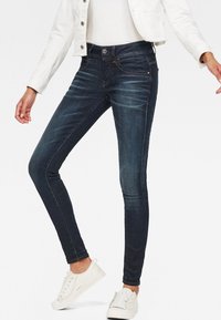 Vaqueros oscuros ajustados de color azul con una textura suave, ligero desgaste, diseño de cinco bolsillos y costuras contrastantes. Combinados con zapatillas blancas.