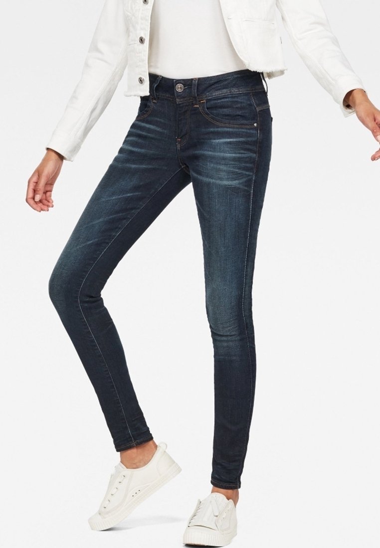 Vaqueros oscuros ajustados de color azul con una textura suave, ligero desgaste, diseño de cinco bolsillos y costuras contrastantes. Combinados con zapatillas blancas.