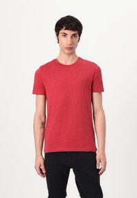 Rotes Baumwoll-T-Shirt mit kurzen Ärmeln und rundem Ausschnitt. Mit einem kleinen Logo auf der Brust. Getragen mit schwarzen Hosen.