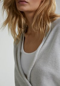 Cardigan tricoté gris clair en matériau doux, avec une coupe décontractée et un motif texturé, superposé à un débardeur blanc.