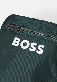 Grøn nylon taske med en struktureret overflade; har et fremtrædende hvidt "BOSS" logo og en mørk lynlåse mekanisme langs den øverste kant.