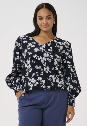 Bloes met bloemenprint in marineblauw met witte en lichtblauwe patronen; V-hals, ballonmouwen en elastische manchetten. Gecombineerd met op maat gemaakte blauwe broeken.