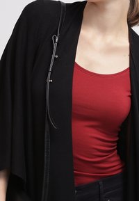 Vrouw in een aansluitende rode tanktop onder een losse zwarte cardigan met een zwarte leren strap over haar schouder.